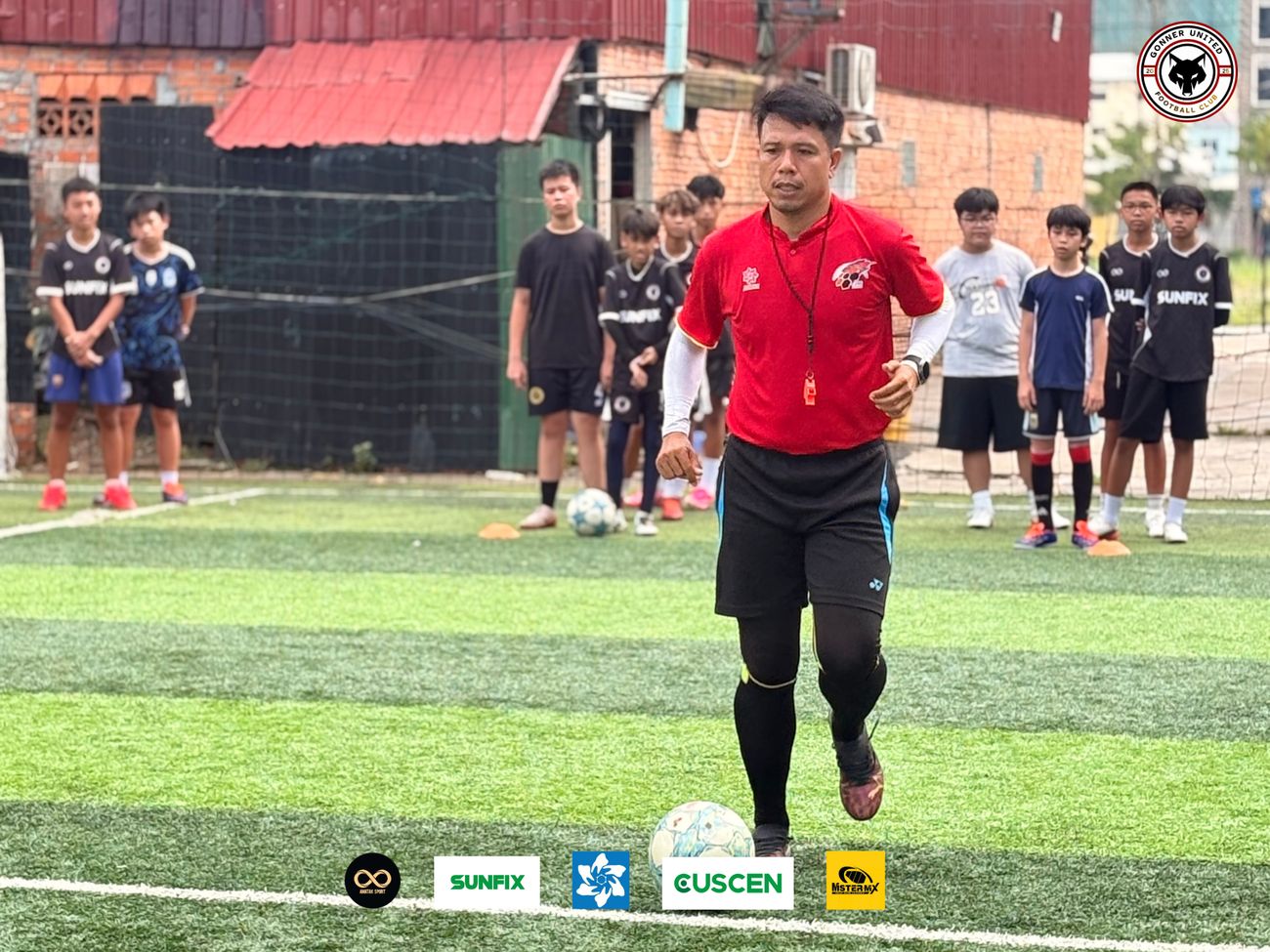 A coach in a red shirt with a whistle runs with a ball while Gonner United FC players watch from behind